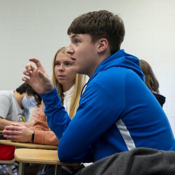 Student seated at desk gesturing with hand, other students in the background