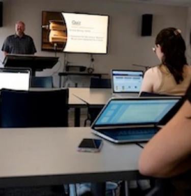 Students seated at laptops in classroom while instructor speaks at podium